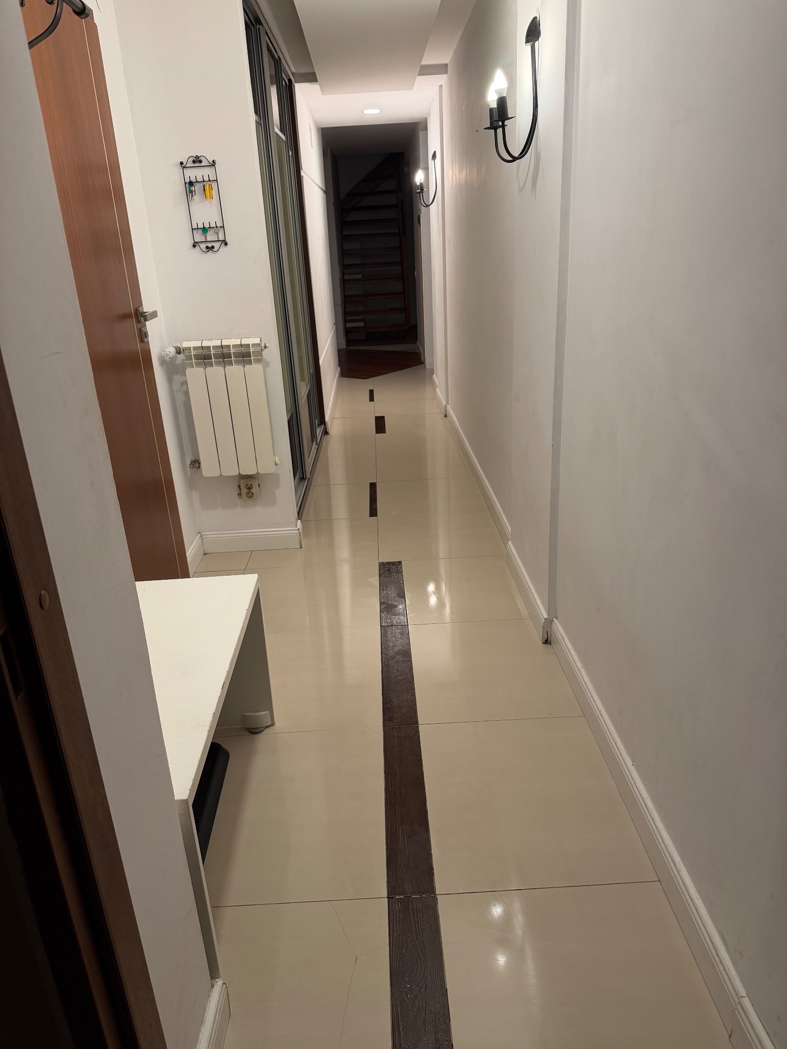 Długi, wąski korytarz z beżowymi płytkami i ciemnobrązowym pasem na środku. Białe ściany, czarne kinkiety, grzejnik i wieszak na klucze. Widoczne schody w głębi.