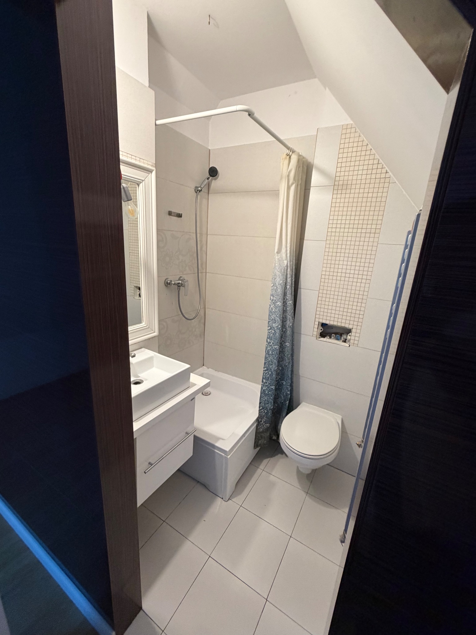 Wykończona łazienka w Elblągu: białe płytki, wanna z prysznicem, umywalka z szafką i toaleta. Uwagę zwraca skośny sufit i minimalistyczny design.