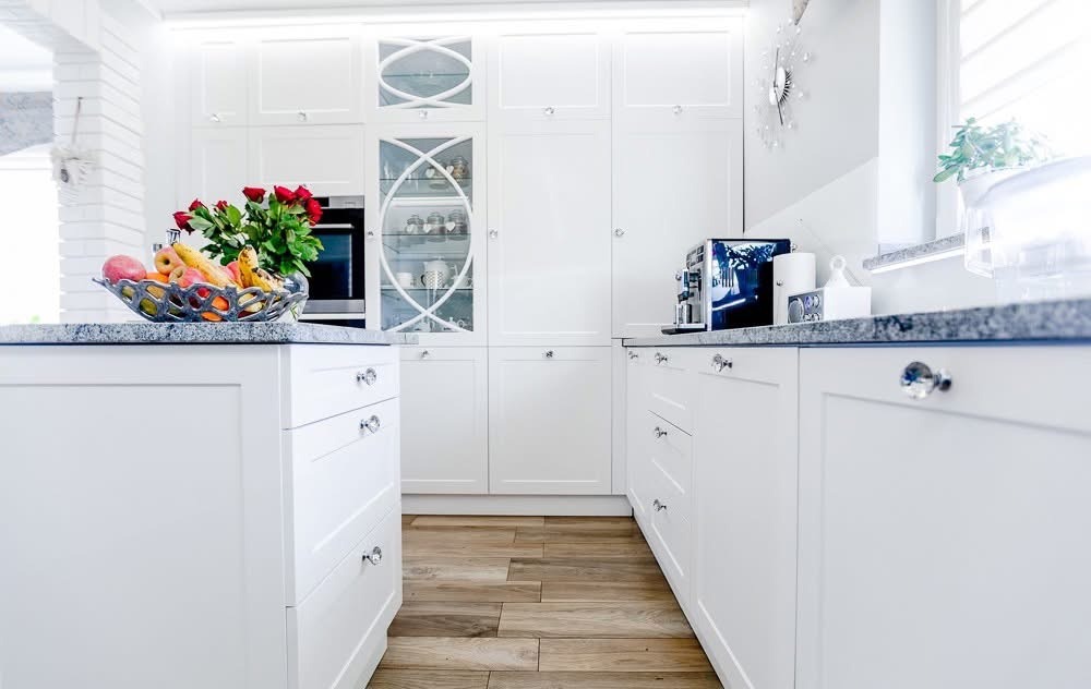 Biała, luksusowa kuchnia z drewnianą podłogą, granitowym blatem i dekoracyjnymi uchwytami. Na blacie misa z owocami i ekspres do kawy. Widoczne przeszklone szafki.