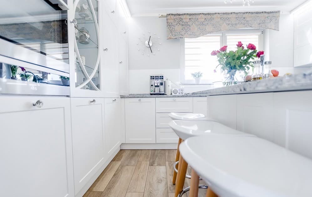 Jasna kuchnia w stylu skandynawskim: białe meble z frezowanymi frontami, drewniana podłoga, kwiaty w wazonie i nowoczesny ekspres do kawy na blacie.