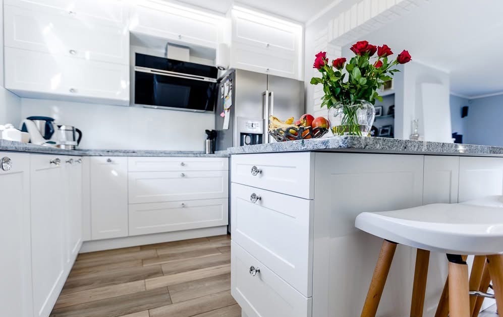 Biała kuchnia z wyspą, blatem z szarego granitu i bukietem czerwonych róż. Nowoczesne meble z frezowanymi frontami i kryształowymi gałkami. Jasne drewniane panele na podłodze.