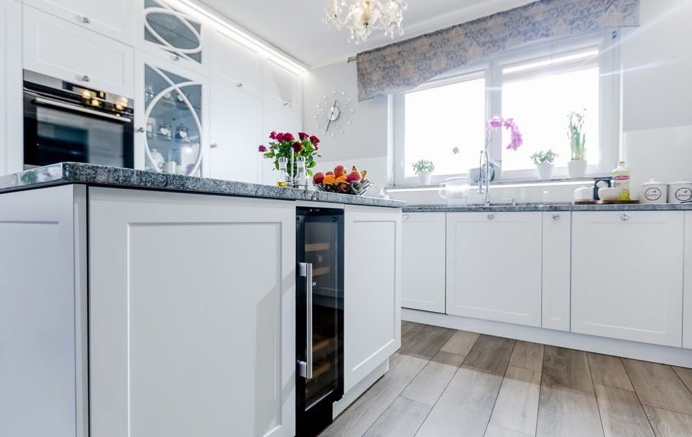 Biała kuchnia z wyspą i wbudowaną chłodziarką na wino. Na blacie granitowym owoce i kwiaty. Okno z roletą rzymską. Parkiet w odcieniach szarości.