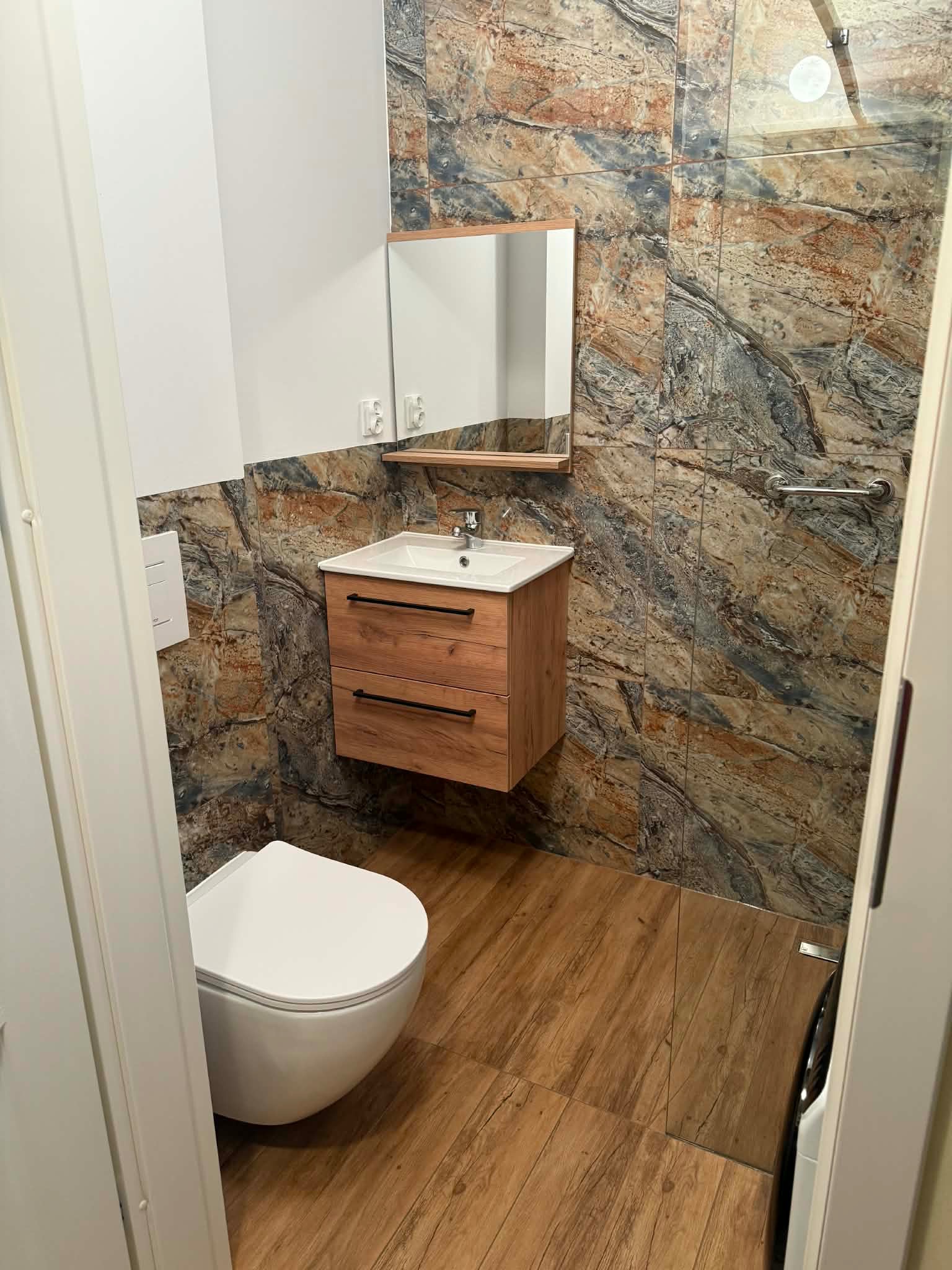 Nowoczesna łazienka po remoncie: toaleta, umywalka z szafką z drewna, lustro i płytki imitujące marmur. Widok zza otwartych drzwi.