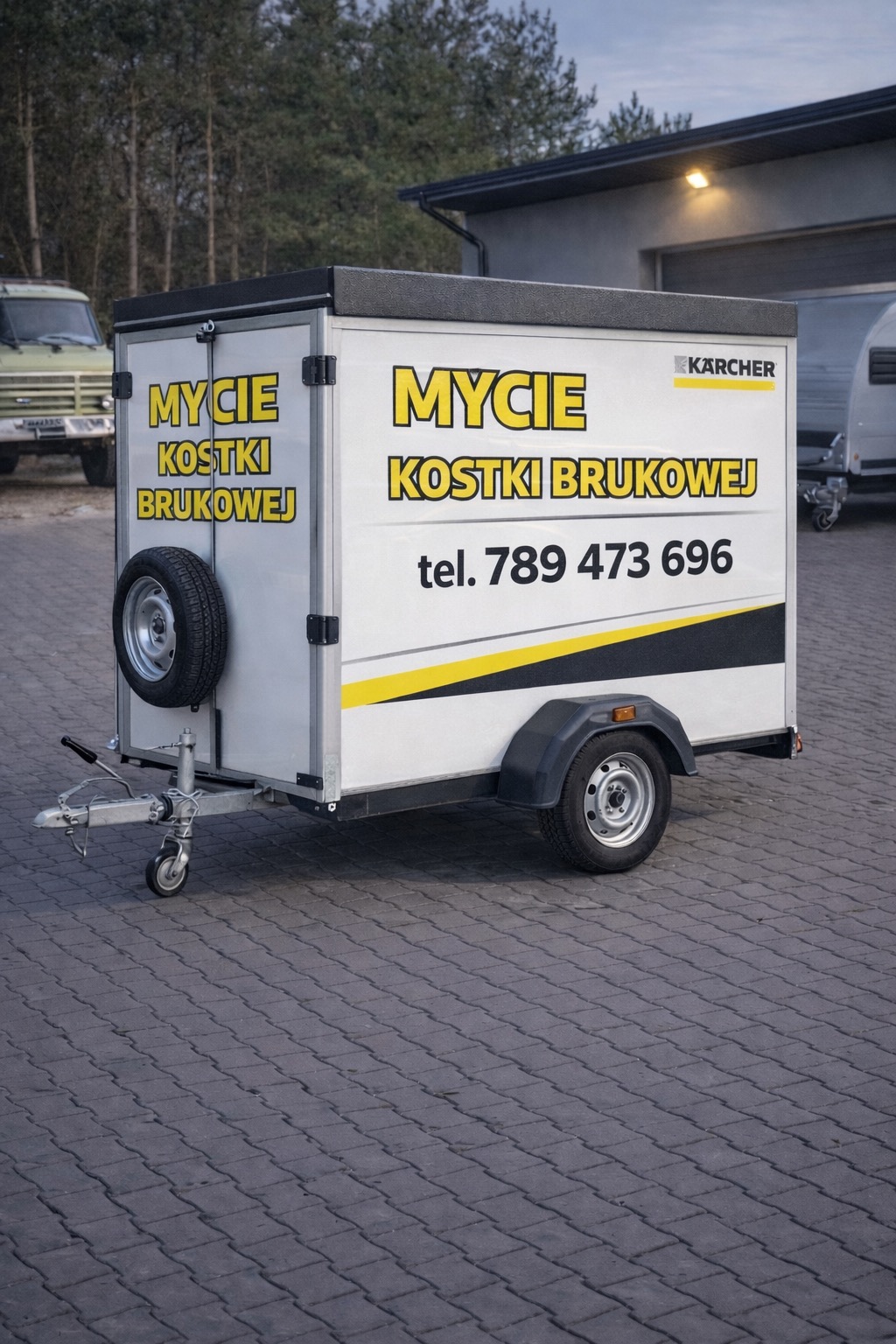 Przyczepa z reklamą firmy Karcher oferującej mycie kostki brukowej, zaparkowana na kostce brukowej. Widoczny numer telefonu kontaktowego.
