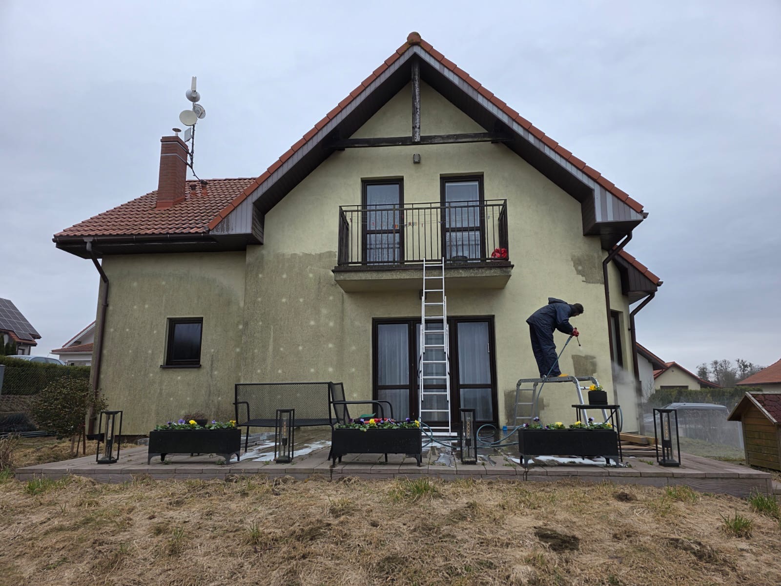 Czyszczenie elewacji domu jednorodzinnego myjką ciśnieniową. Widoczny fragment wyczyszczonej ściany, drabina i pracownik w kombinezonie ochronnym.