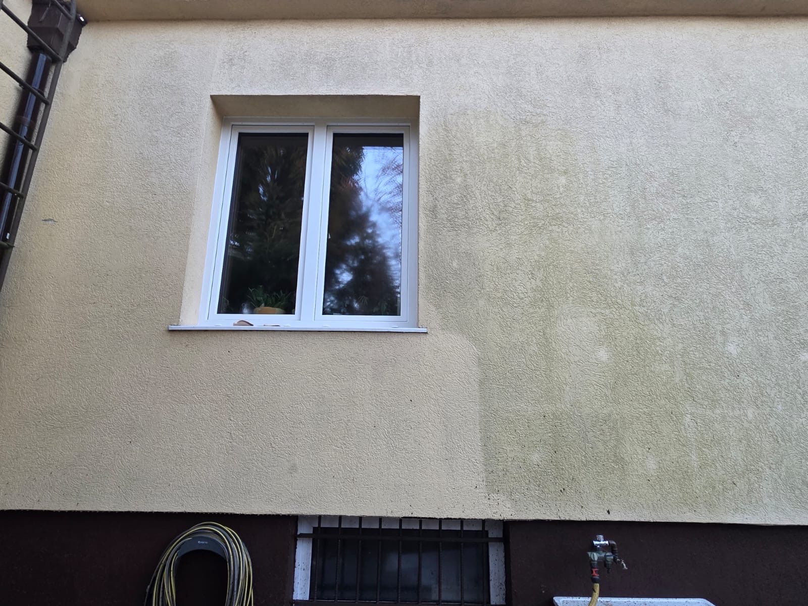 Fragment elewacji budynku z widocznym efektem mycia: czysta powierzchnia kontrastuje z zabrudzoną. Okno z białymi ramami, w tle zieleń. Wąż ogrodowy i kran przy ścianie.