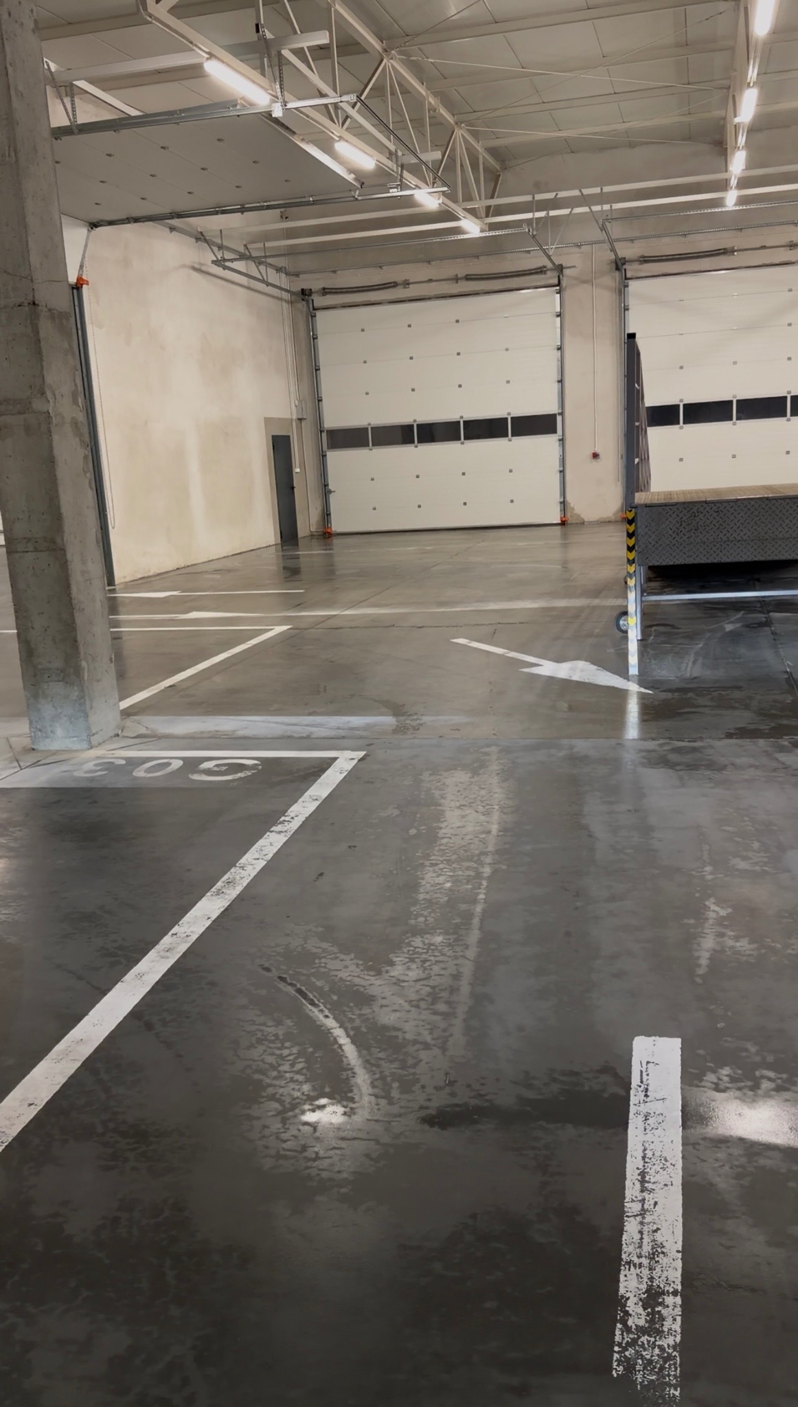 Błyszcząca, mokra posadzka hali magazynowej z białymi liniami wyznaczającymi miejsca parkingowe i strzałkami kierunkowymi. W tle widoczne bramy wjazdowe.