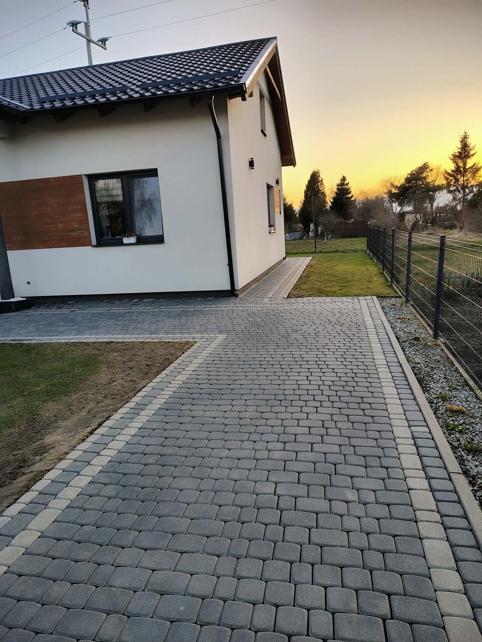 Ułożona kostka brukowa w odcieniach szarości prowadząca do nowoczesnego domu z białymi ścianami i drewnianymi akcentami, widok na ogród i zachodzące słońce.