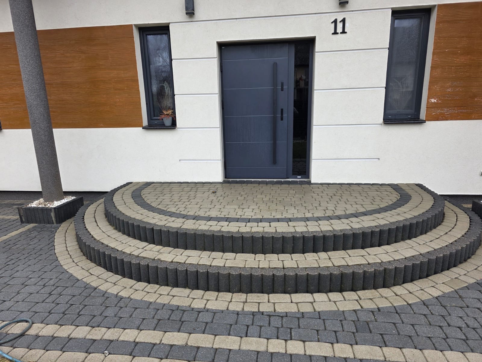 Ułożona kostka brukowa przed domem z numerem 11, tworząca półokrągłe schody wejściowe. Szara, nowoczesna stolarka okienna i drzwi. Elementy drewniane na elewacji.
