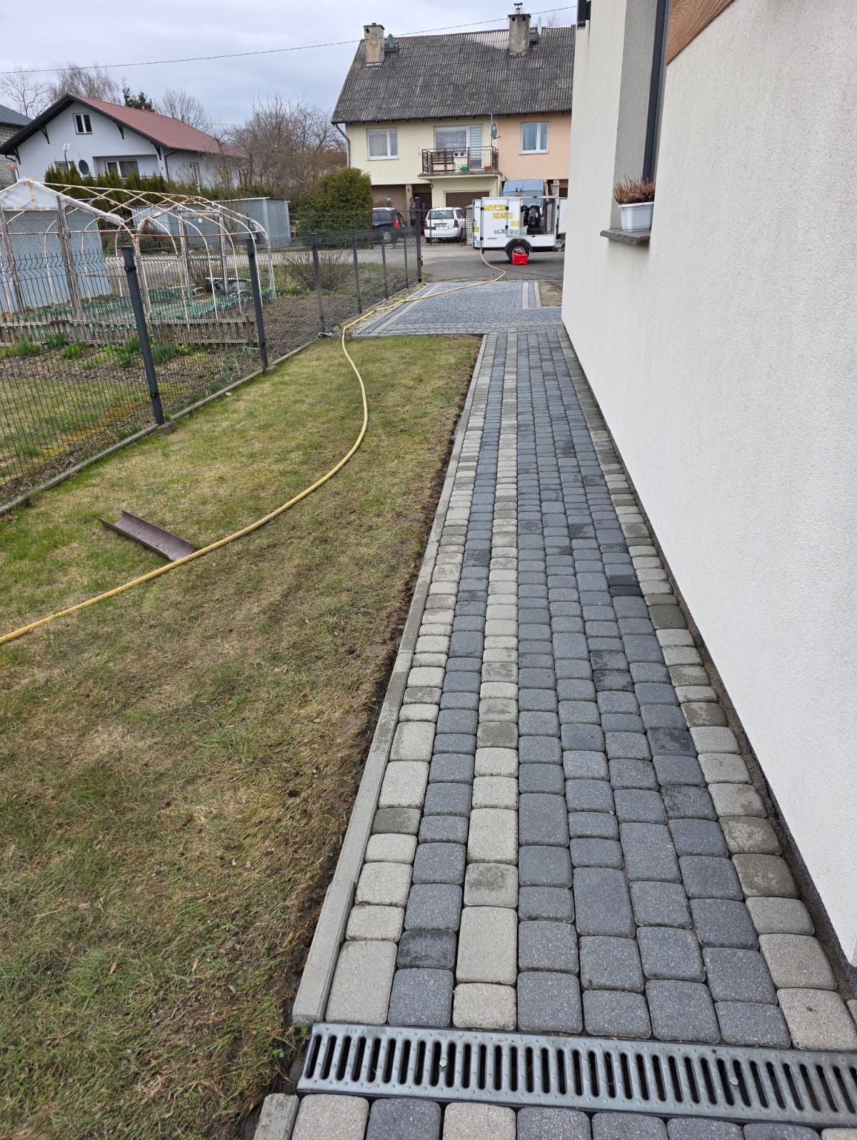 Ułożona kostka brukowa w odcieniach szarości tworzy ścieżkę wzdłuż białej ściany budynku. Widoczny trawnik i żółty wąż ogrodowy. W tle zabudowa jednorodzinna.