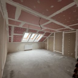 VOSER Sp. z o.o. - Poddasze z oknami dachowymi w trakcie prac. Widoczne płyty na suficie i ścianach, przygotowane do dalszych prac wykończeniowych. Surowy beton na podłodze.