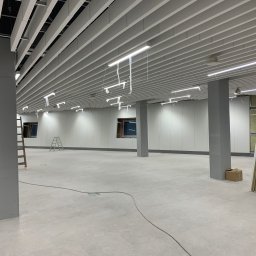 VOSER Sp. z o.o. - Wnętrze biurowe w trakcie prac wykończeniowych: sufit rastrowy z oświetleniem, szare kolumny, białe ściany, drabiny i materiały budowlane. Widok na przestronne pomieszczenie.