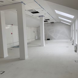VOSER Sp. z o.o. - Jasne, przestronne wnętrze biurowe w trakcie prac wykończeniowych. Widoczne kolumny, okna dachowe i ściany przygotowane do dalszych prac. Podłoga pokryta warstwą wyrównującą.