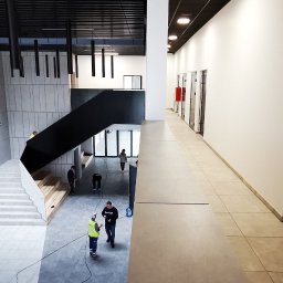 VOSER Sp. z o.o. - Wnętrze biurowca w trakcie prac wykończeniowych: widok na hol z betonową ścianą, schodami i pracownikami w kamizelkach odblaskowych. Minimalistyczny design.