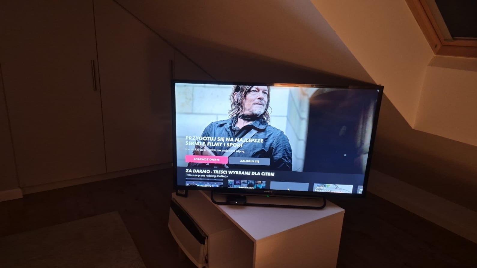 Telewizor Sony Bravia w pokoju na poddaszu, wyświetlający ofertę Canal+ z Normanem Reedusem. Biała szafka RTV i skosy dodają wnętrzu przytulnego charakteru.