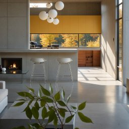 ICON STUDIO ALEKSANDER PAWLUS - Nowoczesne wnętrze domu z betonowymi ścianami, kominkiem i żółtymi szafkami. Widok na jesienny ogród przez duże okno. Minimalistyczne meble i oświetlenie.