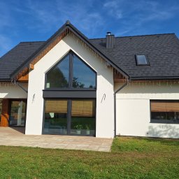 Tarka term Łukasz Tarka - Nowoczesny dom jednorodzinny z białą elewacją, grafitową stolarką i dachem z dachówki. Dekoracyjne lampki zwisają z okapu. Widoczne okno dachowe i trawnik.