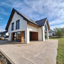 Nowoczesny dom z białymi ścianami i drewnianymi akcentami, z dużym tarasem wyłożonym jasną kostką, otoczony zielonym trawnikiem i błękitnym niebem.