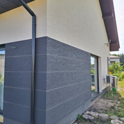 Tarka term Łukasz Tarka - Dwukolorowa elewacja domu z wyraźną fakturą i czarną rynną, widok z boku, z fragmentem okna i klimatyzatora. Tło stanowią domy i zieleń.