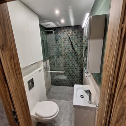 Tarka term Łukasz Tarka - Nowoczesna łazienka po remoncie: biała armatura, kabina prysznicowa z geometrycznymi płytkami w odcieniach zieleni i brązu, szara podłoga, drewniane obramowanie drzwi.