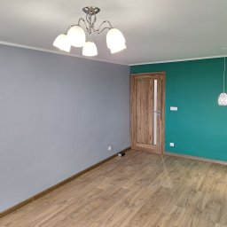 Tarka term Łukasz Tarka - Wnętrze po malowaniu: szara ściana z klimatyzatorem kontrastuje z zieloną ścianą z drewnianymi drzwiami i designerskimi lampami. Na podłodze panele.