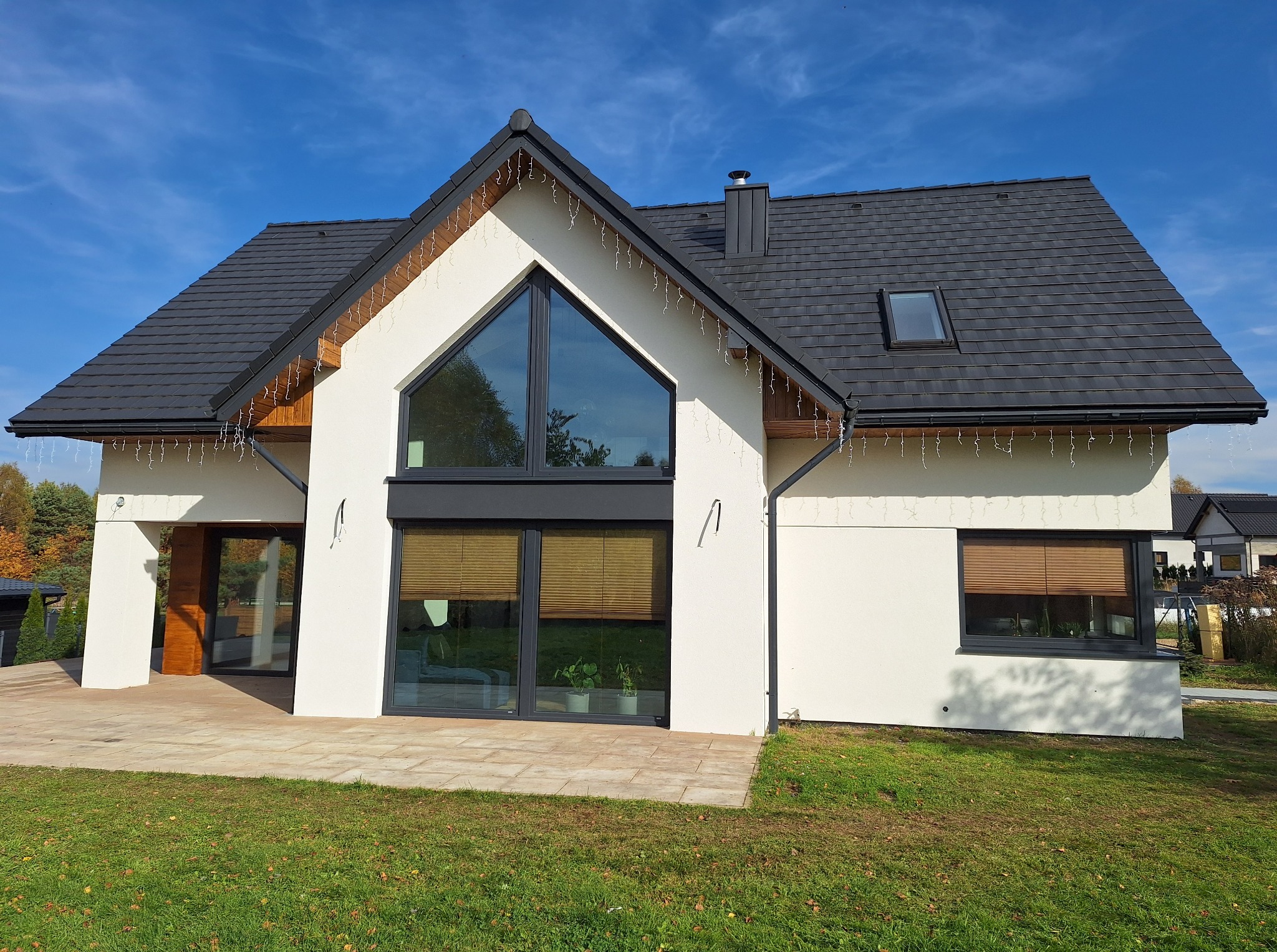 Nowoczesny dom jednorodzinny z białą elewacją, grafitową stolarką i dachem z dachówki. Dekoracyjne lampki zwisają z okapu. Widoczne okno dachowe i trawnik.