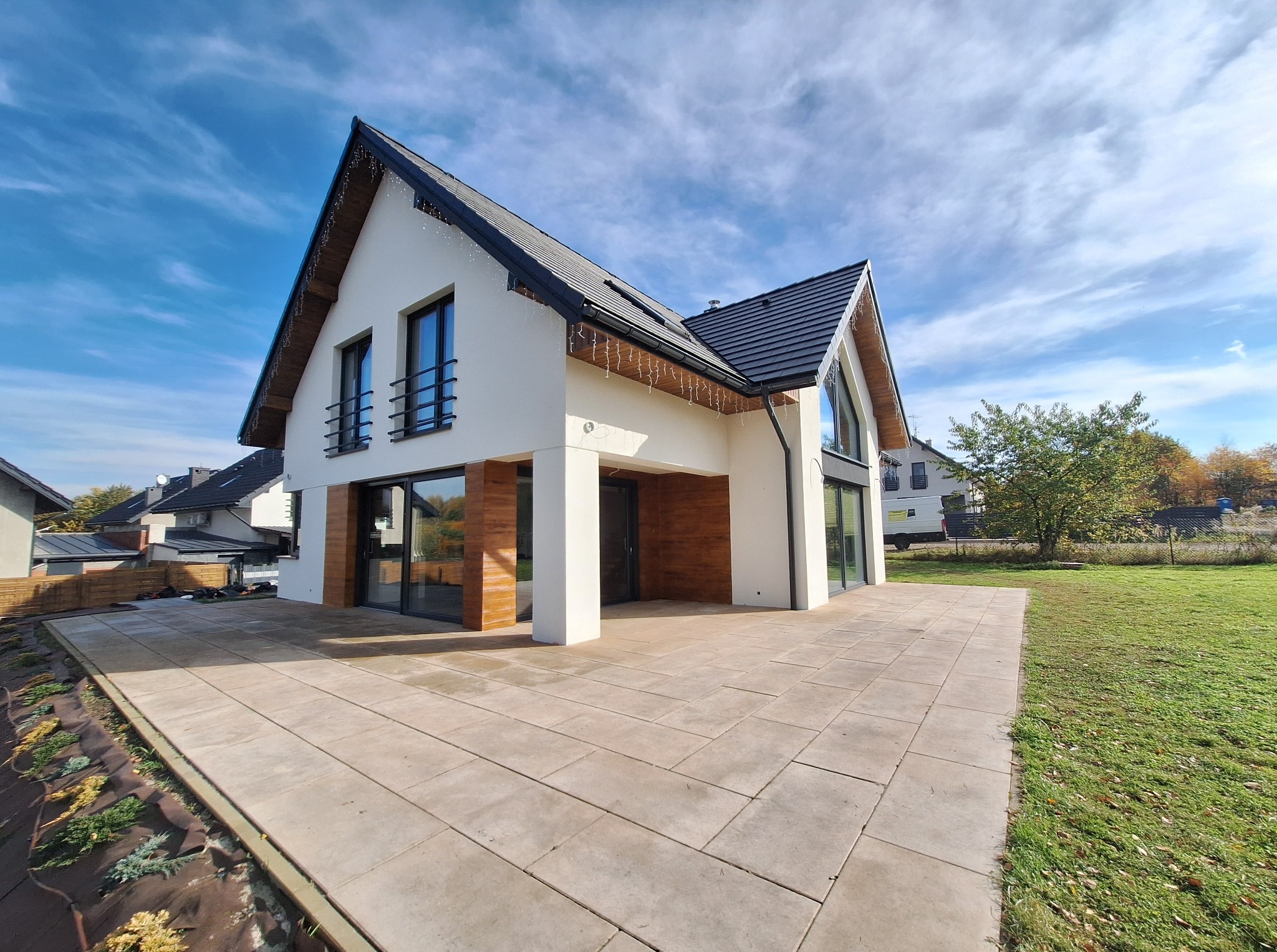 Nowoczesny dom z białymi ścianami i drewnianymi akcentami, z dużym tarasem wyłożonym jasną kostką, otoczony zielonym trawnikiem i błękitnym niebem.