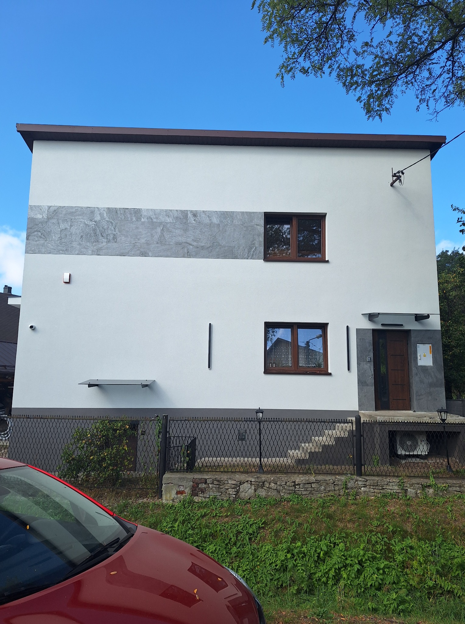 Dwukondygnacyjny dom z białą elewacją, szarym pasem dekoracyjnym i brązowymi oknami. Widoczny fragment czerwonego samochodu i ogrodzenie z metalowej siatki.