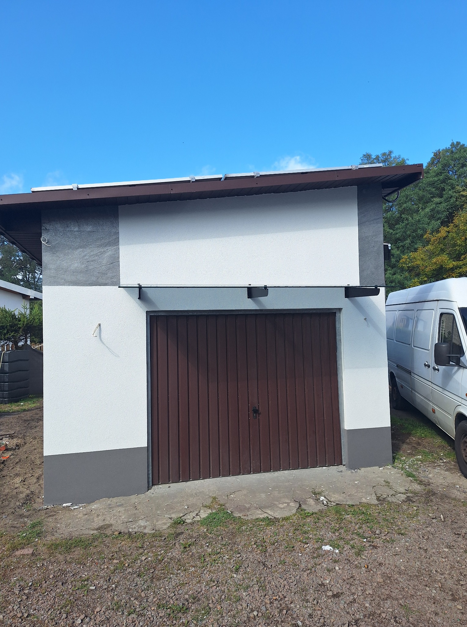 Wykończony garaż z brązową bramą i świeżo pomalowaną elewacją w odcieniach bieli i szarości, z zaparkowanym obok białym busem.