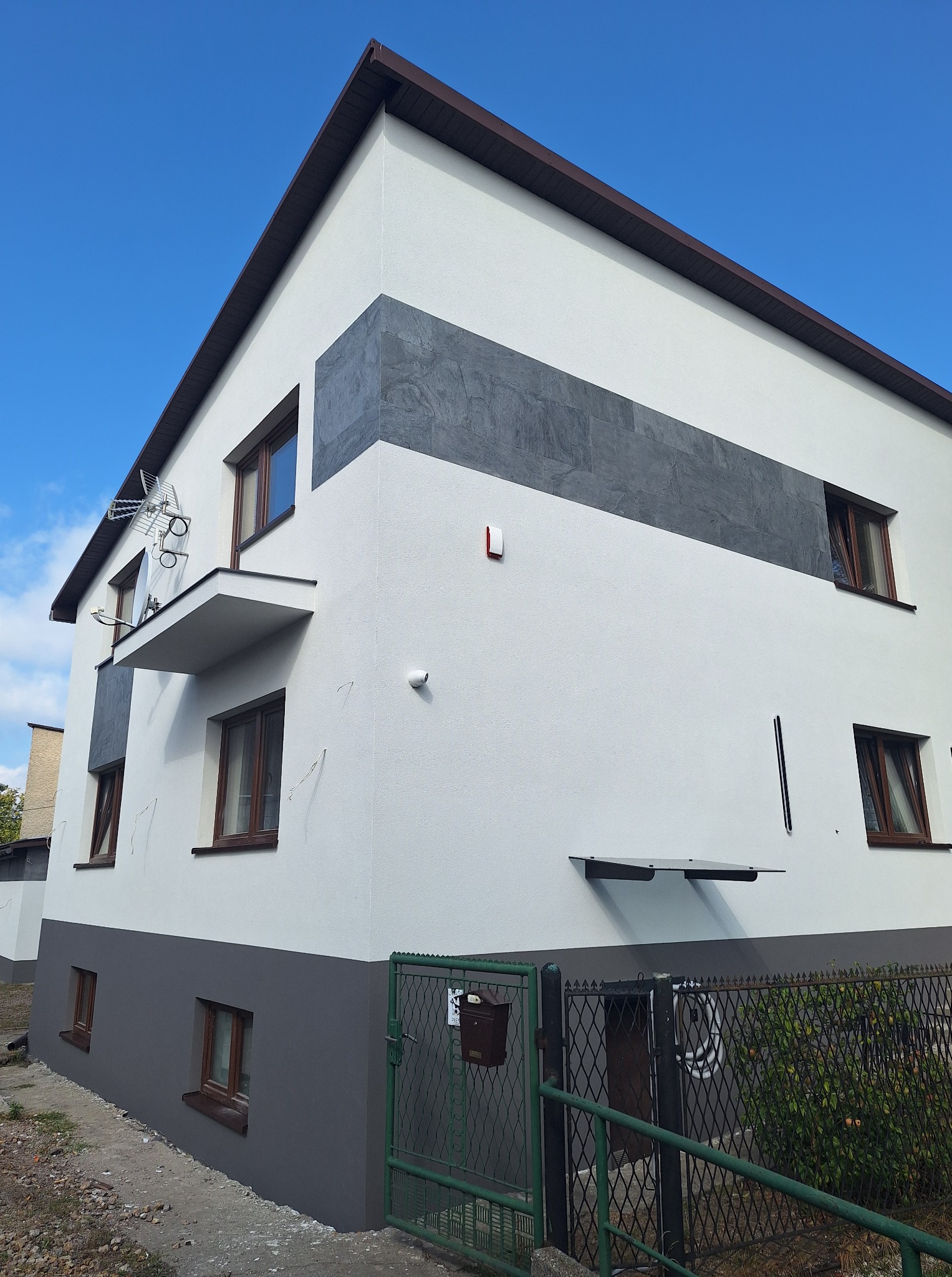 Dwukondygnacyjny dom po renowacji elewacji z szarym pasem dekoracyjnym, brązowymi oknami i zielonym ogrodzeniem, sfotografowany w słoneczny dzień.