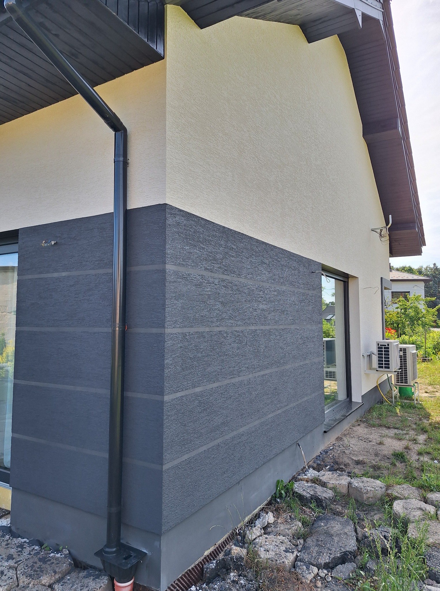 Dwukolorowa elewacja domu z wyraźną fakturą i czarną rynną, widok z boku, z fragmentem okna i klimatyzatora. Tło stanowią domy i zieleń.