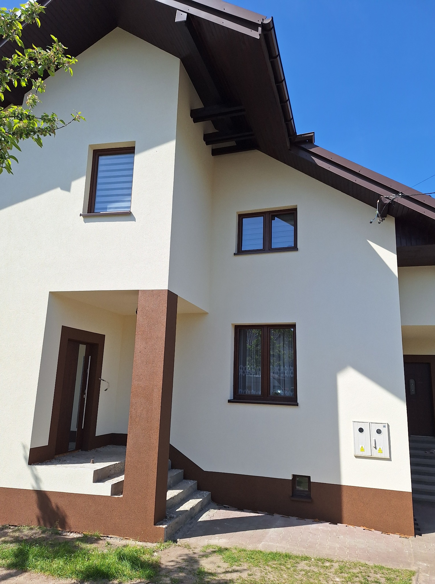 Dwukondygnacyjny dom z beżową elewacją i brązowymi akcentami. Widoczne okna, dach i schody wejściowe. Elewacja świeżo po renowacji w słoneczny dzień.