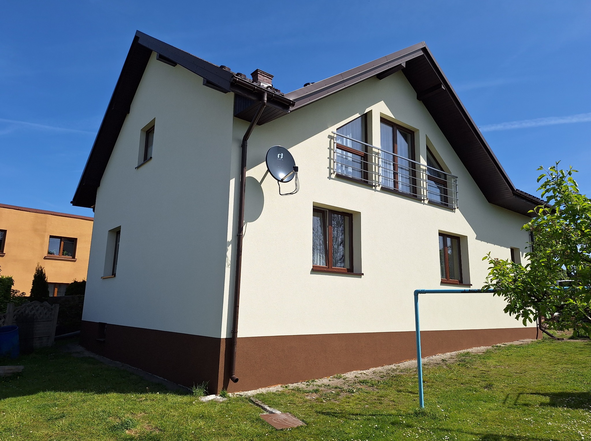 Ocieplony dom jednorodzinny z beżową elewacją i brązowym cokołem, widoczny z boku w słoneczny dzień. Na ścianie zamontowana antena satelitarna.