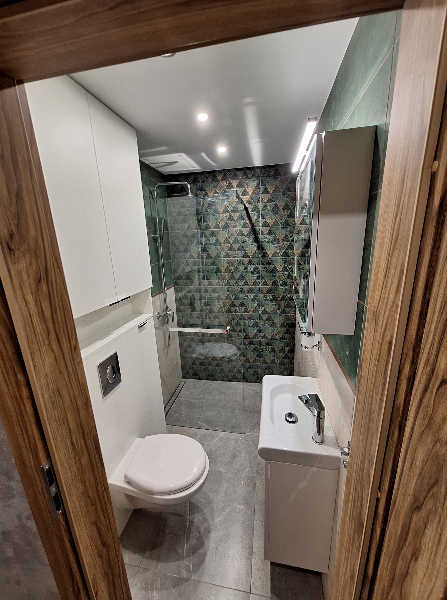 Nowoczesna łazienka po remoncie: biała armatura, kabina prysznicowa z geometrycznymi płytkami w odcieniach zieleni i brązu, szara podłoga, drewniane obramowanie drzwi.