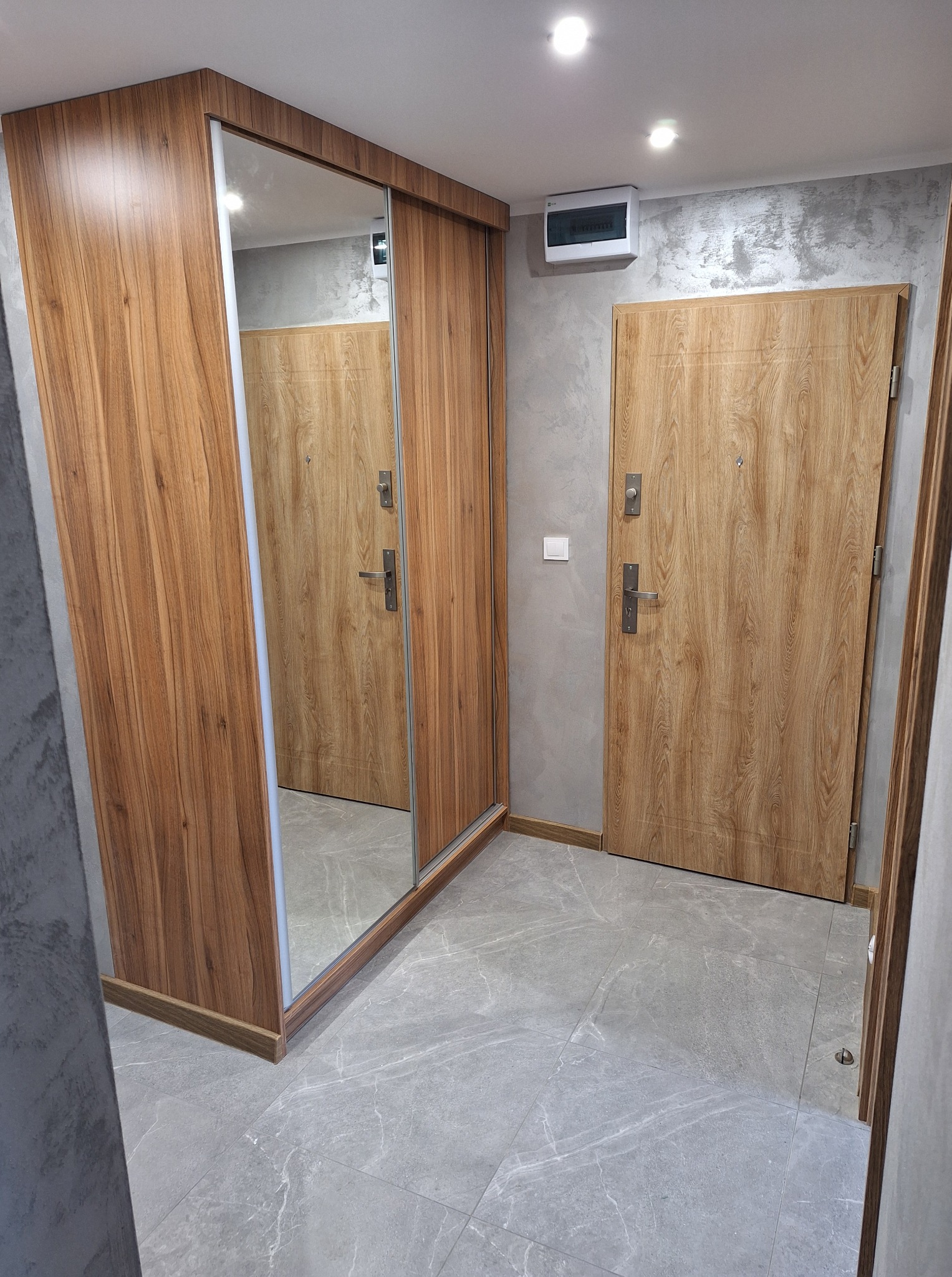 Nowoczesny przedpokój z drewnianą szafą przesuwną z lustrem i drzwiami wejściowymi o podobnym wykończeniu, szare ściany i podłoga z płytek imitujących marmur.