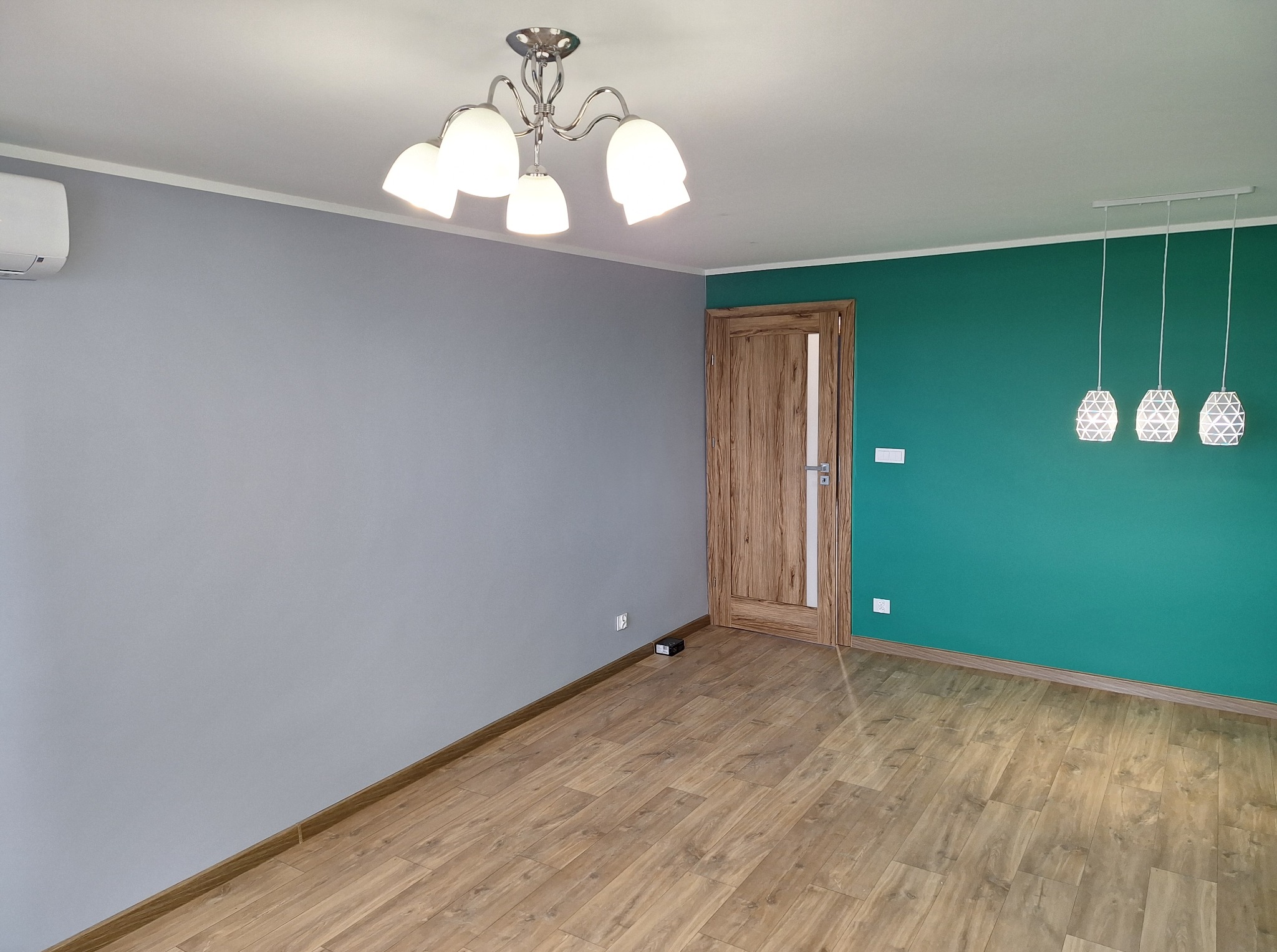 Wnętrze po malowaniu: szara ściana z klimatyzatorem kontrastuje z zieloną ścianą z drewnianymi drzwiami i designerskimi lampami. Na podłodze panele.