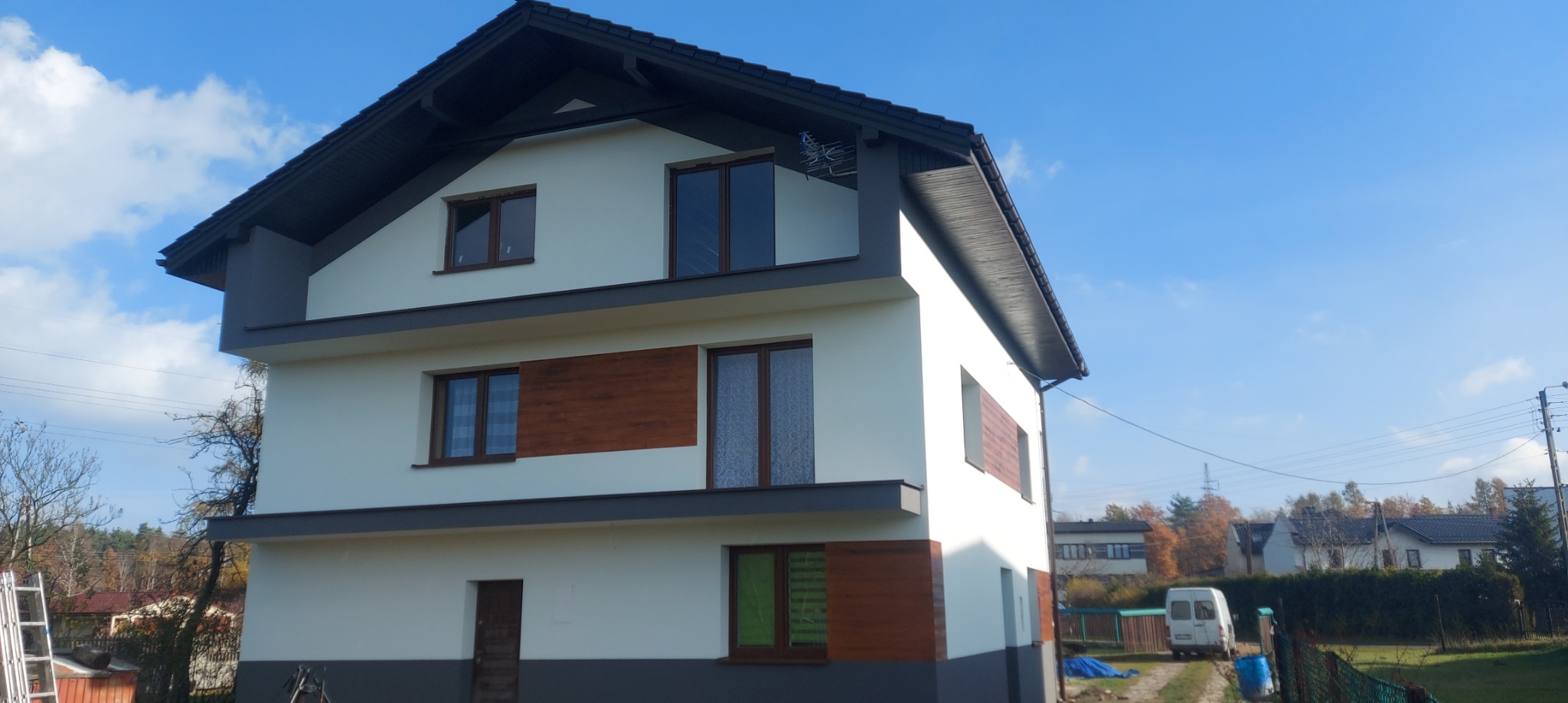 Trzykondygnacyjny dom po renowacji elewacji, z elementami dekoracyjnymi imitującymi drewno, ciemnoszarą podmurówką i dachem, na tle błękitnego nieba.
