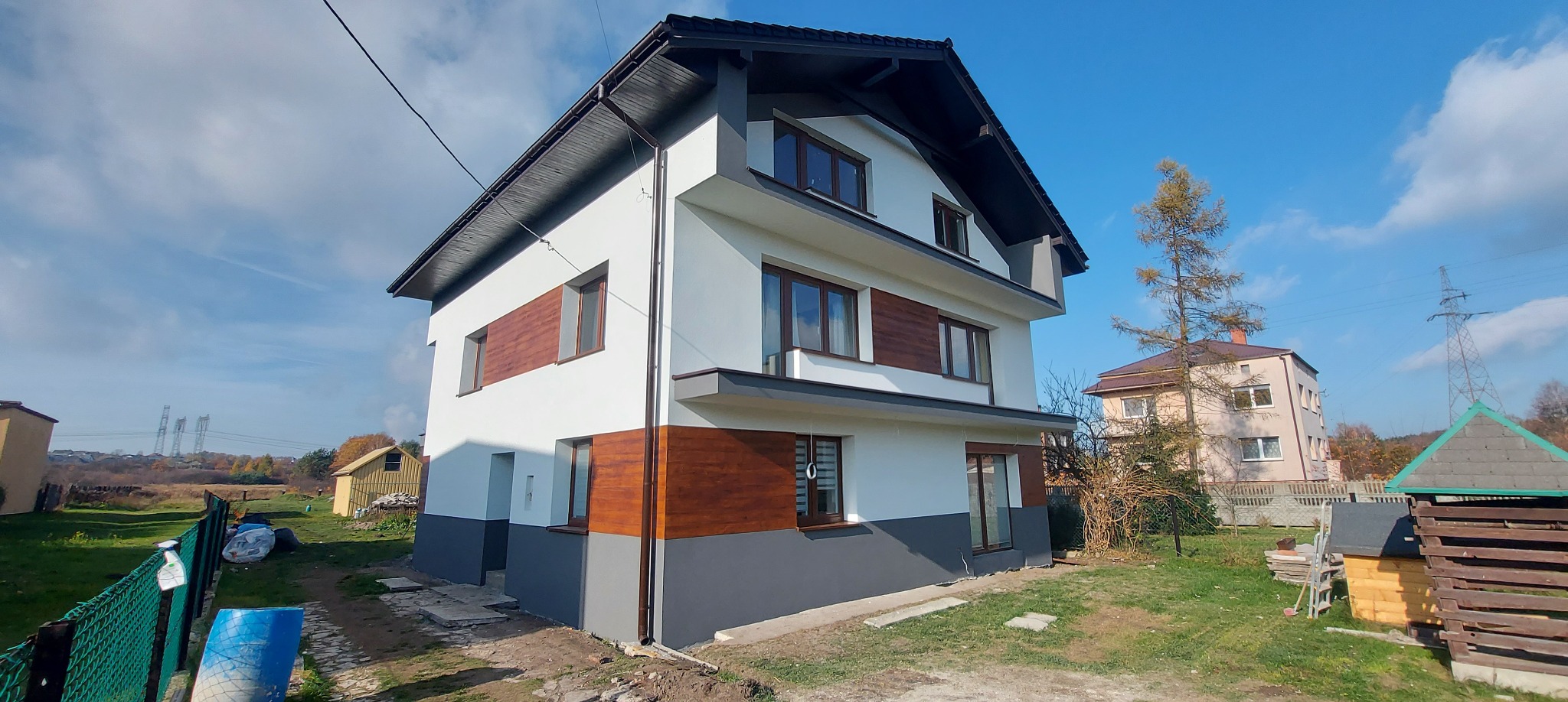 Dwukondygnacyjny dom po renowacji elewacji z elementami dekoracyjnymi imitującymi drewno. Widoczne detale wykończenia i świeże malowanie. Błękitne niebo w tle.