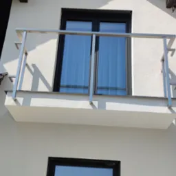 Balkon z balustradą ze stali nierdzewnej, widok z dołu, na tle jasnej elewacji domu i fragmentu dachu z czarną dachówką. Minimalistyczny design i nowoczesne wykończenie.