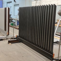 Dziedzic Team - Warsztatowa ekspozycja dwóch segmentów ogrodzenia lamelowego: ciemnoszarego i wielokolorowego, zamocowanych tymczasowo do podłoża. W tle sprzęt spawalniczy i elementy konstrukcyjne.