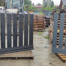 Dziedzic Team - Dwa segmenty nowoczesnego ogrodzenia panelowego: czarny, mokry od deszczu, i szary, ustawione na paletach w magazynie z widocznym traktorem i materiałami budowlanymi.