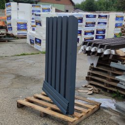 Dziedzic Team - Elementy ogrodzenia panelowego w kolorze grafitowym, ułożone na palecie, czekają na montaż. W tle materiały budowlane i budynek.