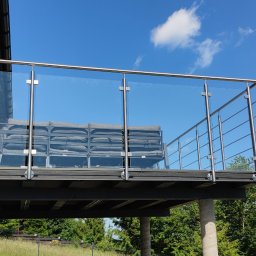 Dziedzic Team - Nowoczesny balkon z balustradą ze stali nierdzewnej i szkła, widok z dołu na tle błękitnego nieba. Minimalistyczny design, metalowe elementy i szklane panele.