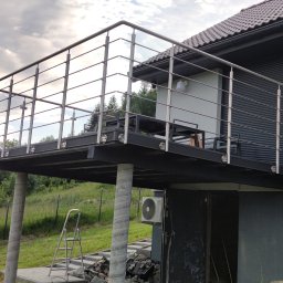 Dziedzic Team - Nowoczesny balkon z balustradą nierdzewną i szklanymi panelami, wsparty na betonowych kolumnach, z widokiem na zalesione wzgórza w tle.