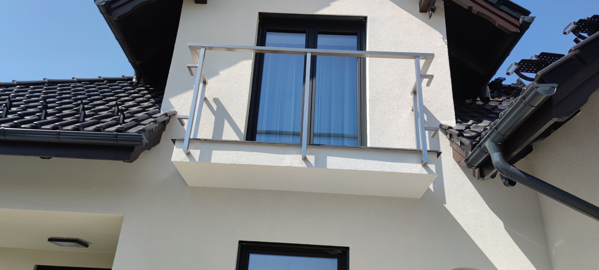 Balkon z balustradą ze stali nierdzewnej, widok z dołu, na tle jasnej elewacji domu i fragmentu dachu z czarną dachówką. Minimalistyczny design i nowoczesne wykończenie.