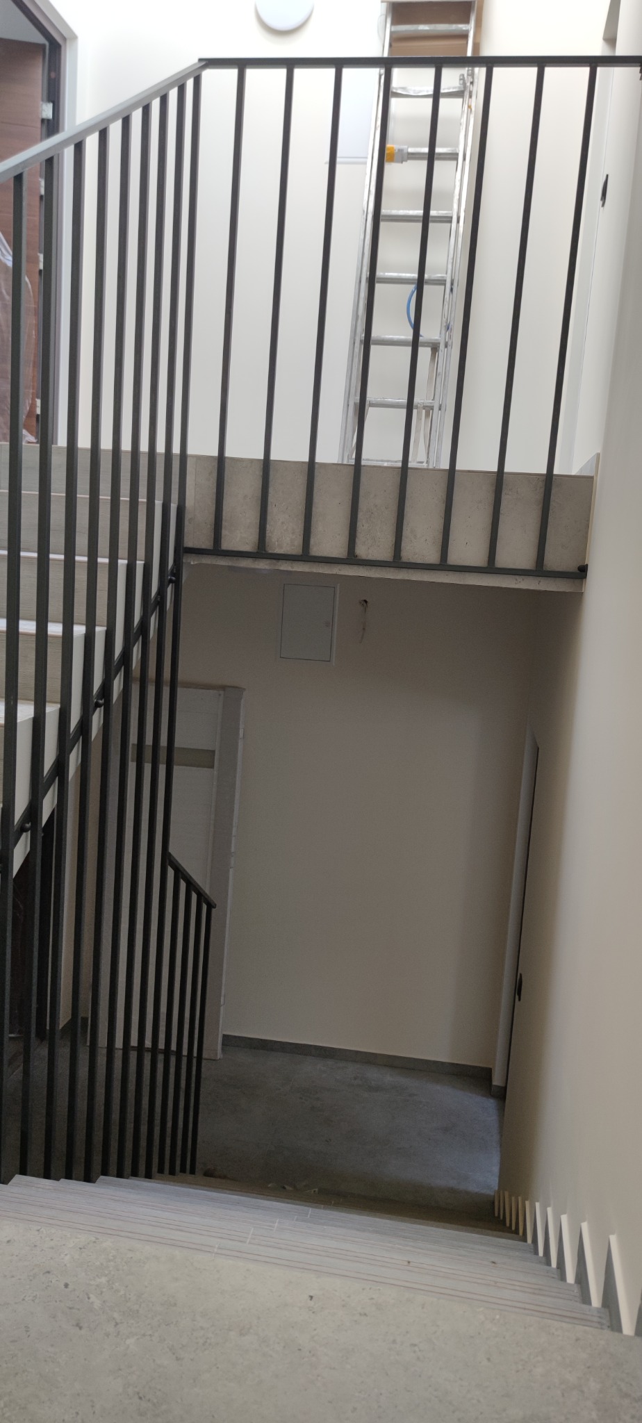 Nowoczesne schody betonowe z czarną metalową balustradą w jasnym wnętrzu klatki schodowej. W tle widoczna drabina oparta o ścianę.