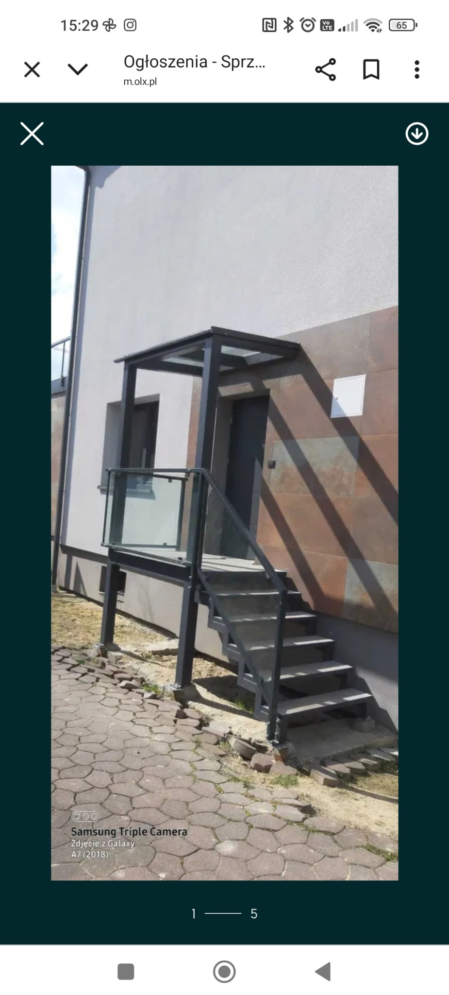 Metalowe schody zewnętrzne z przeszkloną balustradą i zadaszeniem, prowadzące do drzwi wejściowych budynku. Szara kolorystyka konstrukcji na tle jasnej elewacji.