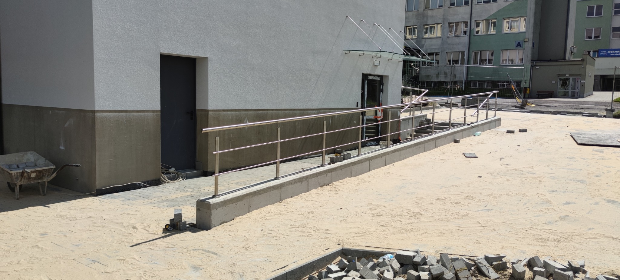 Zewnętrzne, metalowe balustrady przy schodach wejściowych do budynku, z widocznymi elementami placu budowy i kostką brukową. Jasne oświetlenie.