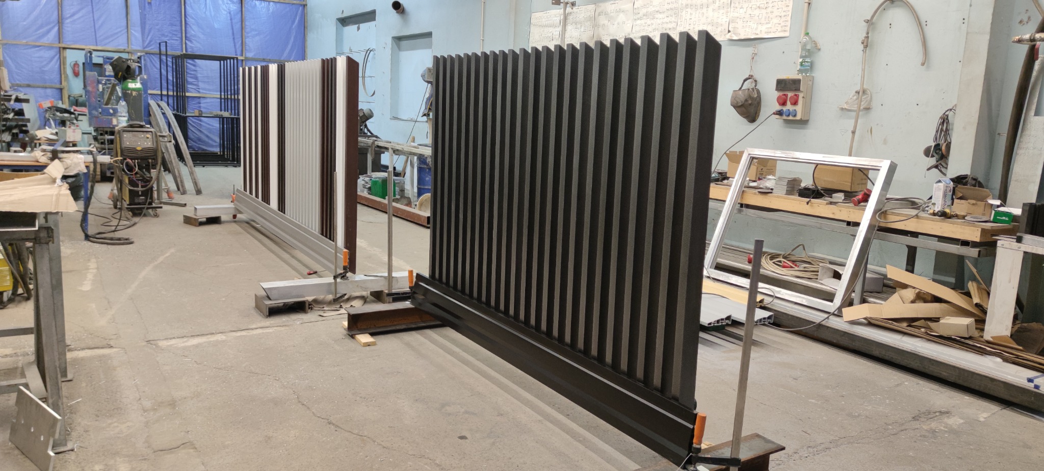 Warsztatowa ekspozycja dwóch segmentów ogrodzenia lamelowego: ciemnoszarego i wielokolorowego, zamocowanych tymczasowo do podłoża. W tle sprzęt spawalniczy i elementy konstrukcyjne.