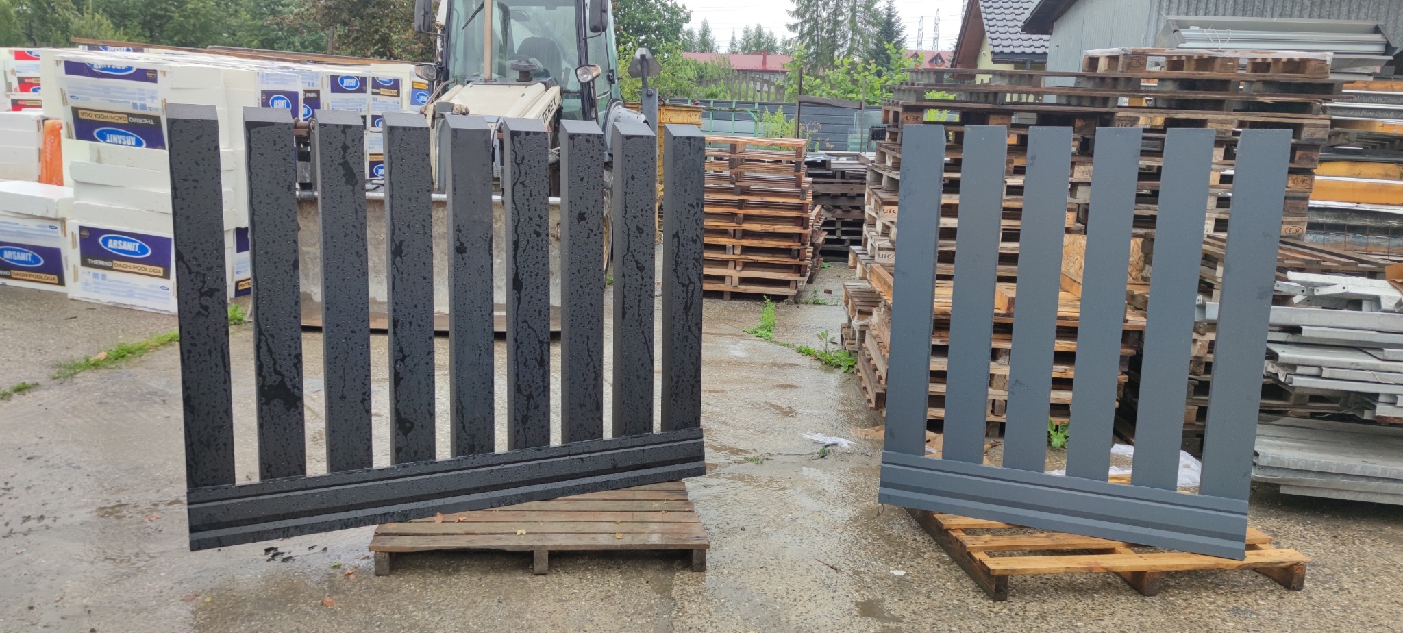 Dwa segmenty nowoczesnego ogrodzenia panelowego: czarny, mokry od deszczu, i szary, ustawione na paletach w magazynie z widocznym traktorem i materiałami budowlanymi.